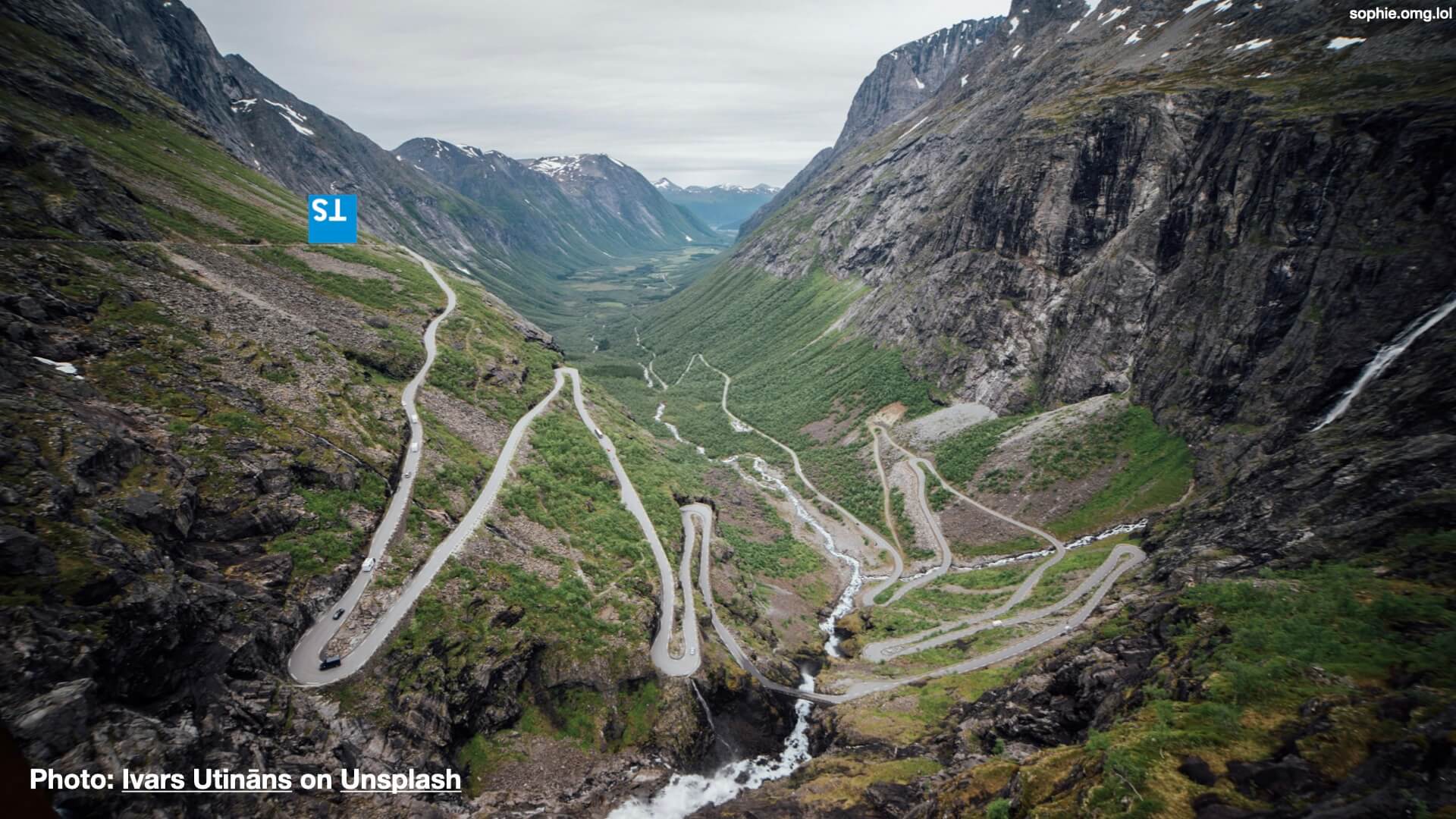 A photograph of a very twisty mountain road surrounded by luscious green and dramatic dark rock cliffs. The Typescript logo is upside down at the top of the road.